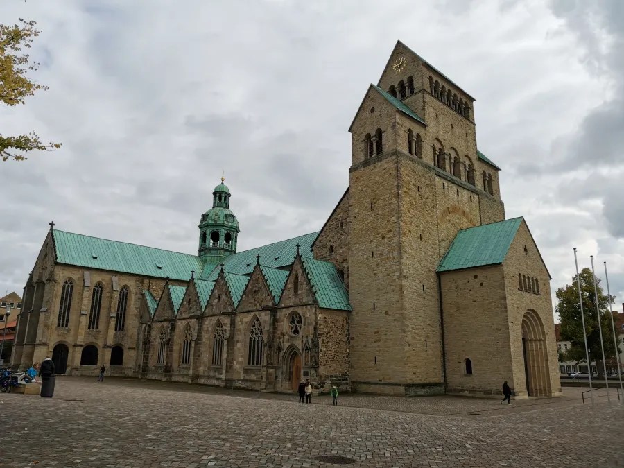 A view of a historic church building with a distinctive green roof and stone facade, situated in an open square under cloudy skies.