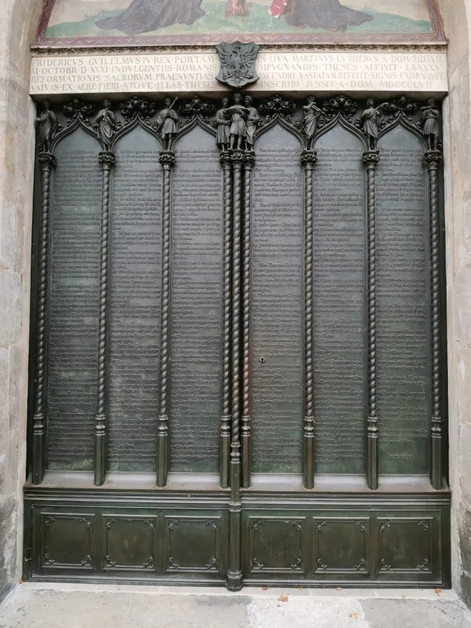 Eine kunstvoll gestaltete Türe mit reliefartigen Verzierungen und Schriftzügen, die an Martin Luthers 95 Thesen erinnert.