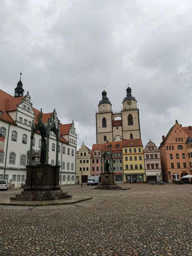 Der Marktplatz in Eisleben mit historischen Gebäuden und zwei Statuen, darunter ein Denkmal von Martin Luther, unter einem bewölkten Himmel.