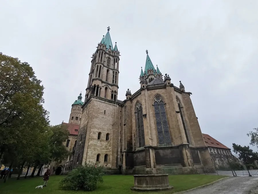 Ansicht eines historischen Gebäudes mit hohen Türmen und grünem Dach, umgeben von Bäumen und einer Wiese.