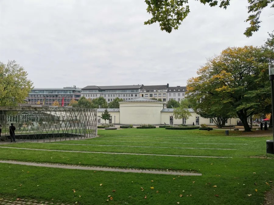 Blick auf eine gepflegte Wiese mit einer modernen Glas- und Stahlstruktur sowie Bäumen vor einem städtischen Hintergrund in Aachen unter bewölktem Himmel.