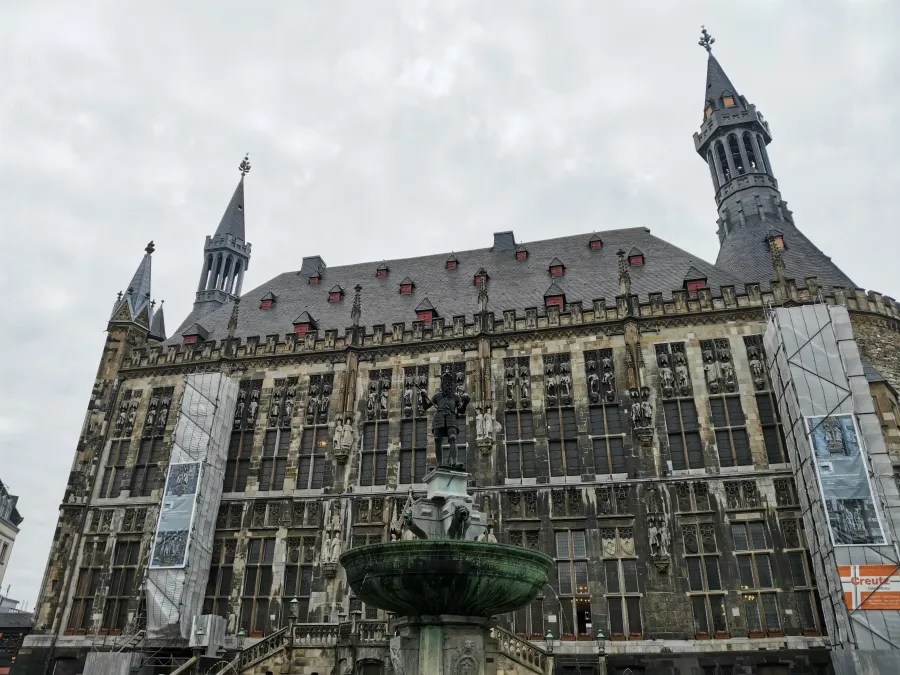 Ansicht des Aachener Rathauses mit einer Brunnenstatue im Vordergrund und grauem Himmel.