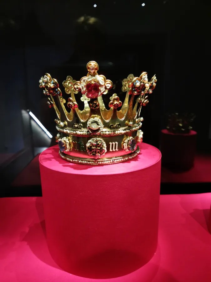 Eine kunstvoll verzierte Krone aus Gold und edelsteinen, auf einem roten Podest präsentiert, im Museum.