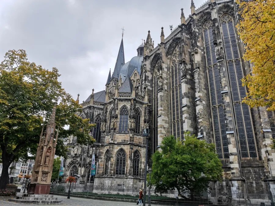 Der Aachener Dom, ein Meisterwerk der karolingischen Renaissance-Architektur, ist von Bäumen umgeben und zeigt seine beeindruckende Fassade unter einem bewölkten Himmel.