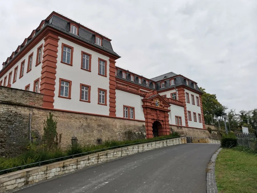 Blick auf ein historisches Gebäude in Mainz mit roter Fassade und zahlreichen Fenstern, umgeben von einer Einfahrt und grüner Landschaft.