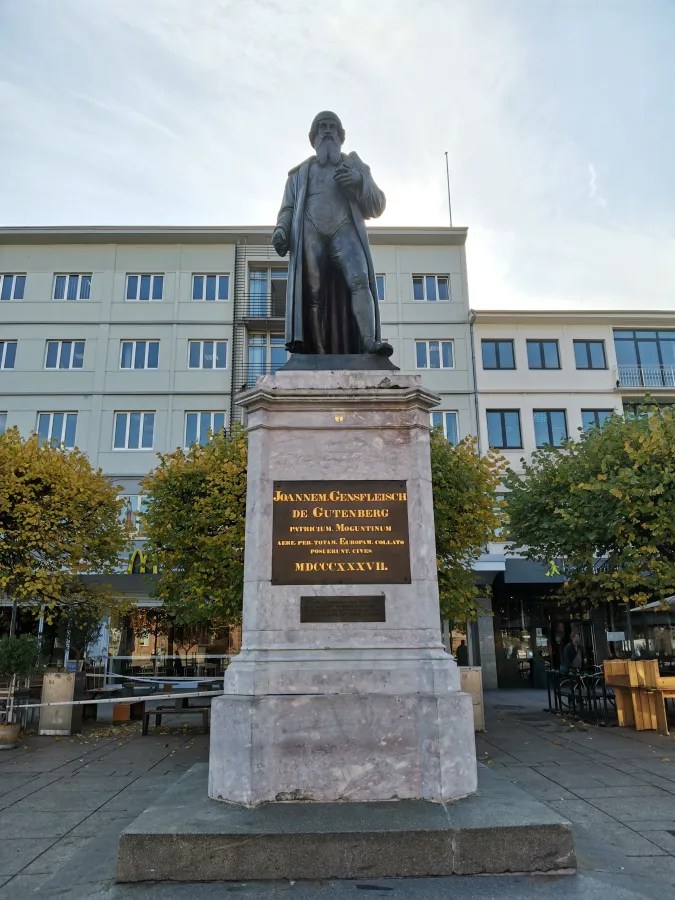 Statue von Johannes Gutenberg auf einem Sockel in Mainz, umgeben von Bäumen und modernen Gebäuden.
