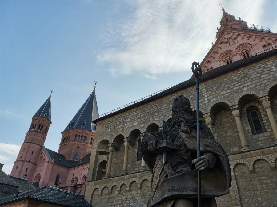 Bronzeskulptur eines heiligen Mannes im Vordergrund, im Hintergrund sind Turmspitzen und Fassaden einer alten Kirche in Mainz zu sehen.