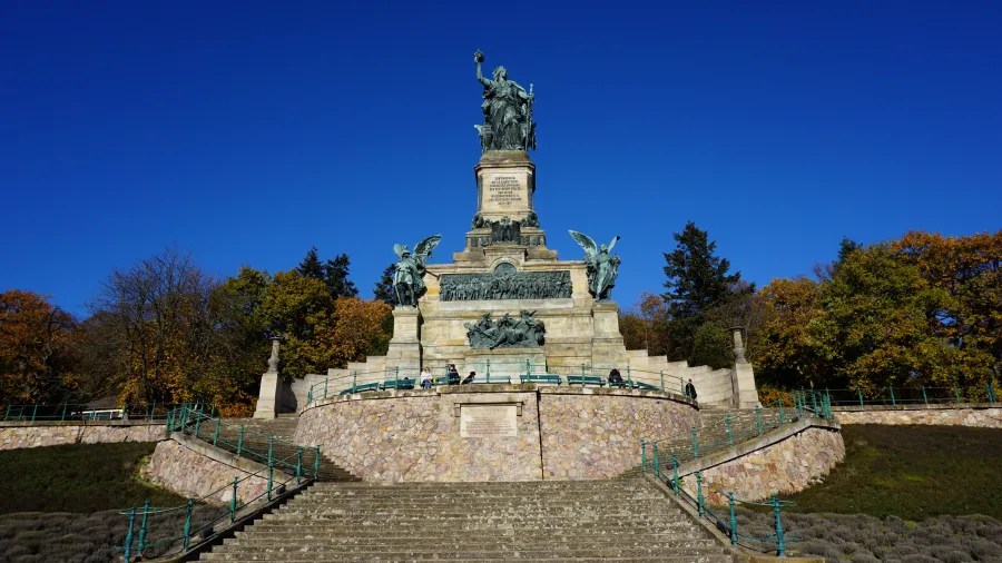 Ein historisches Denkmal mit einer Statue an der Spitze, umgeben von Bäumen im Herbst, unter einem klaren blauen Himmel.