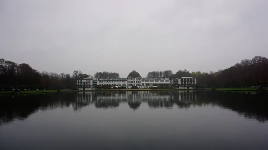 Ein nebliges Herbstbild eines historischen Gebäudes, das sich ruhig im Wasser spiegelt, umgeben von einer grünen Parklandschaft und grauem Himmel.