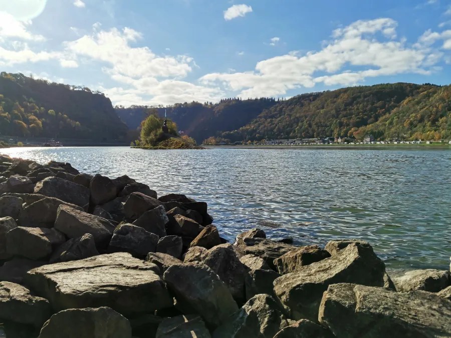 Ausblick auf den Rhein mit Felsen im Vordergrund und bewaldeten Hügeln im Hintergrund, unter einem klaren blauen Himmel mit einigen Wolken.