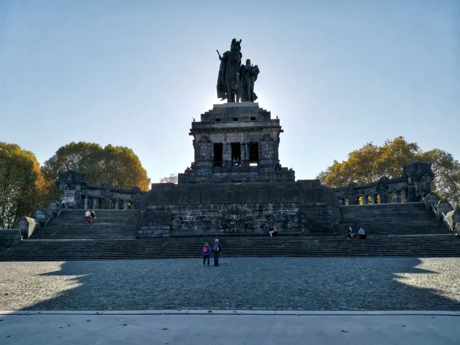 Statue des Deutschen Ecks mit dem Reiterdenkmal im Hintergrund, umgeben von herbstlichen Bäumen und Menschen, die Treppen hinaufsteigen.