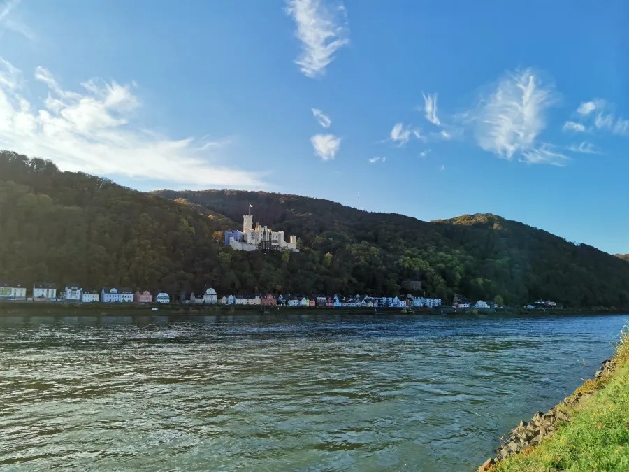 Blick auf eine Burg, die auf einem Hügel über den Rhein thront, umgeben von grünen Wäldern und bunten Häusern am Flussufer unter einem blauen Himmel mit einigen Wolken.