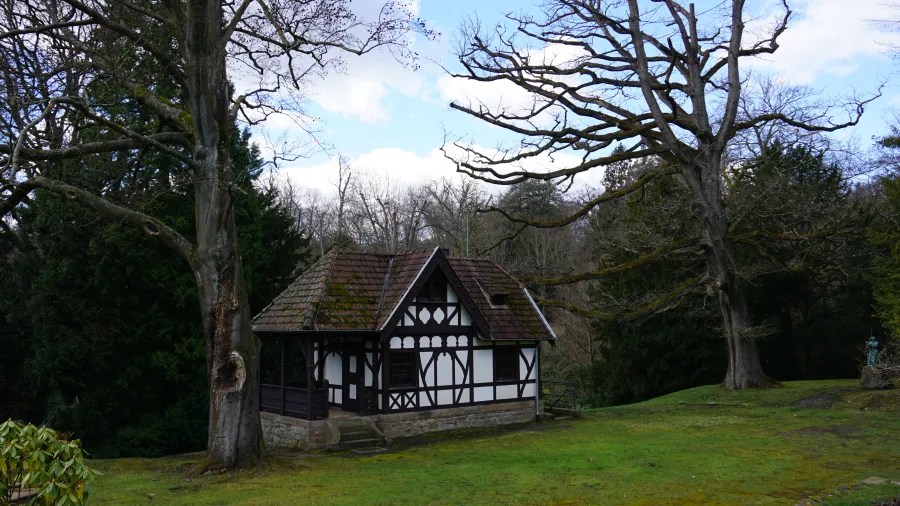 Ein traditionelles Fachwerkhaus steht in einer grünen Wiese, umgeben von großen Bäumen und einer ruhigen Landschaft. Der Himmel ist teilweise bewölkt.