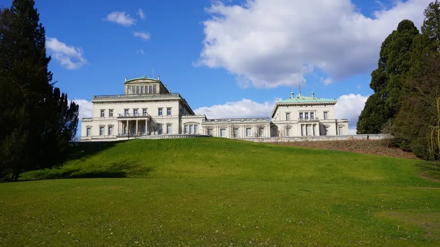 Villa Hügel, ein imposantes Gebäude auf einem grünen Hügel, umgeben von Bäumen und Wiesen unter einem blauen Himmel.