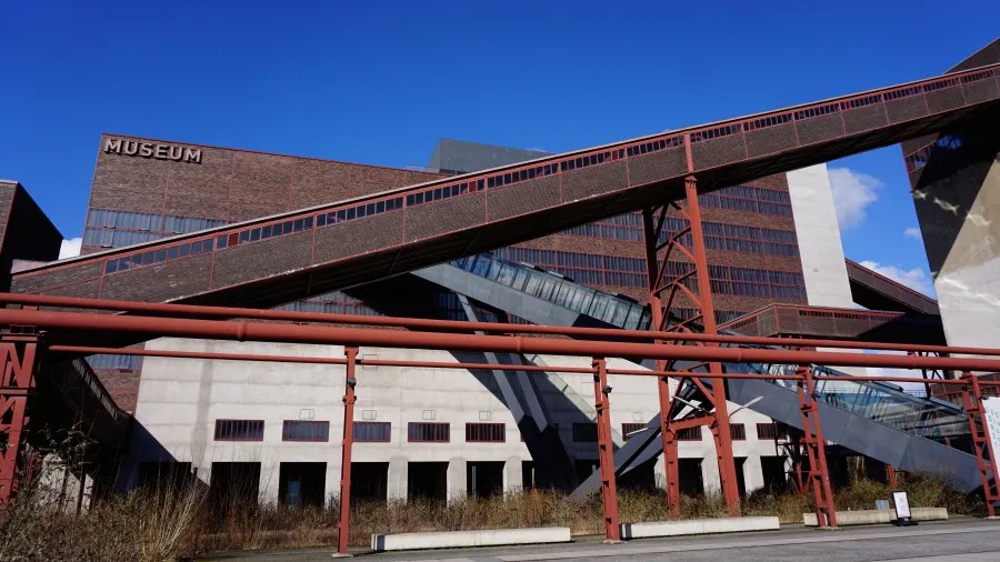 Das Industriedenkmal Zeche Zollverein, inklusive des Museumsgebäudes mit modernen architektonischen Elementen und einer Kombination aus rotem Stahl und Glas, unter blauem Himmel.