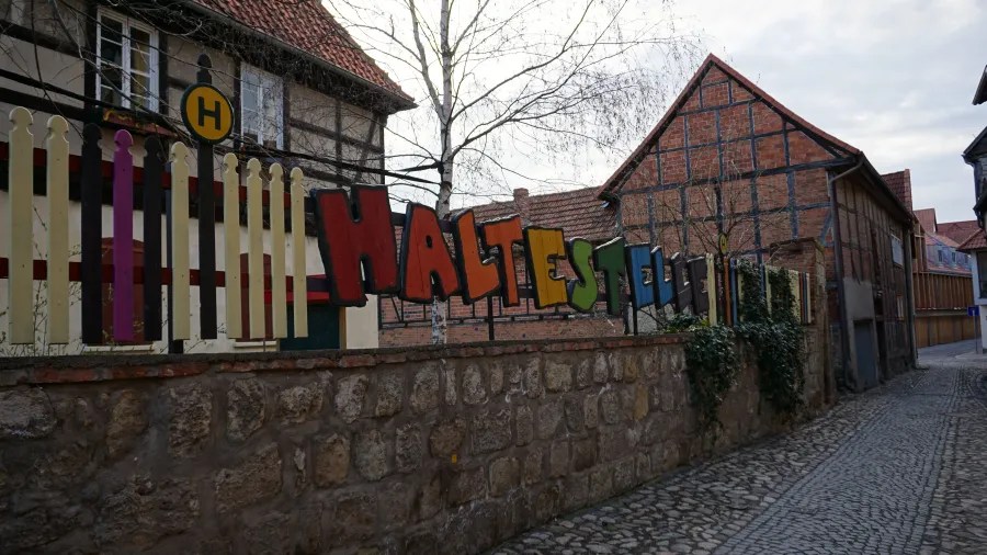 Bunte Buchstaben am Zaun mit der Aufschrift 'Haltestelle' vor einem alten Fachwerkhaus in einer Stadtstraße.