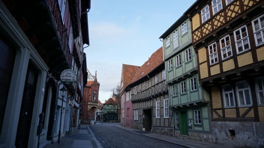 A picturesque street with traditional half-timbered houses in various colors, including green, pink, and yellow, under a clear blue sky.