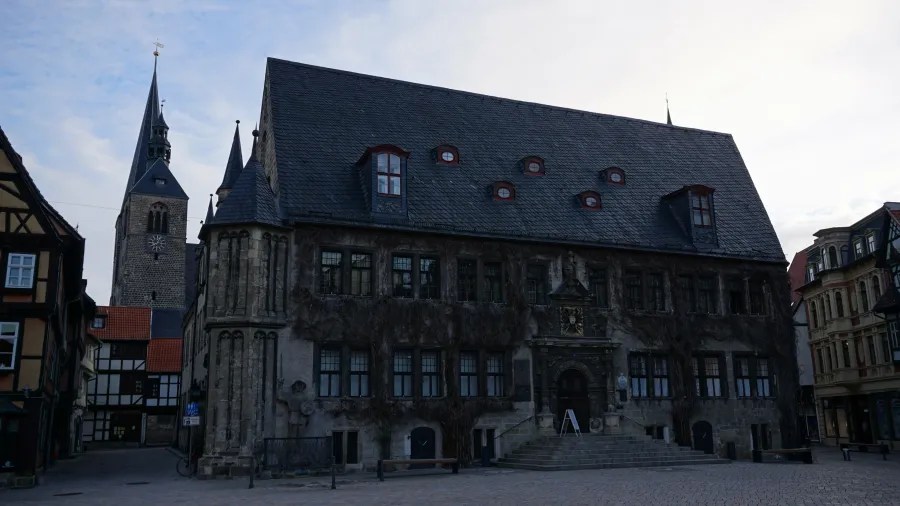Historisches Gebäude mit schiefergedecktem Dach und verwitterter Fassade, umgeben von alten Häusern und einem Kirchturm im Hintergrund.