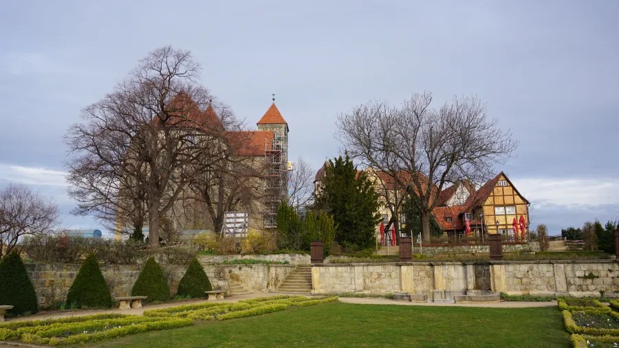 A scenic view of a historic castle and a traditional house surrounded by trees and a garden, with cloudy skies in the background.