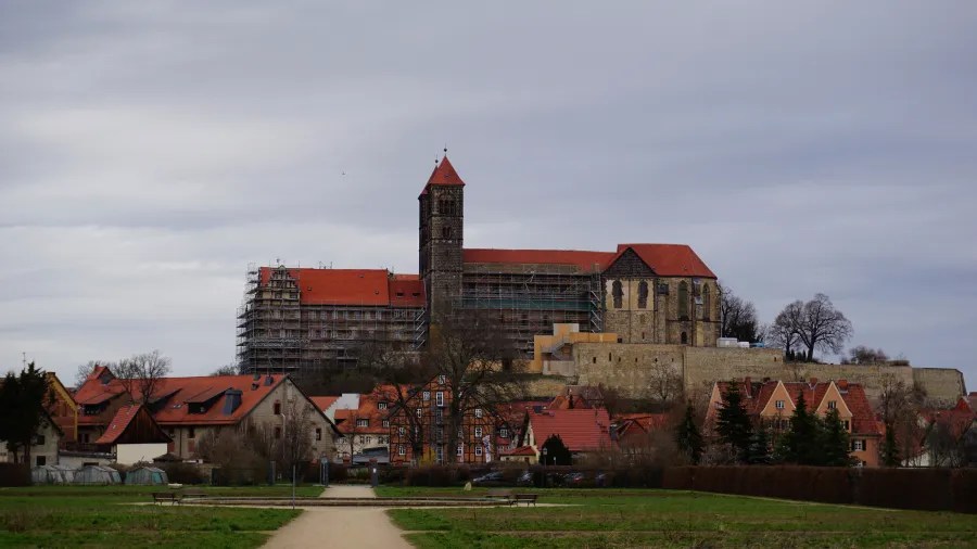 A view of a historical building under restoration, surrounded by houses with red roofs and greenery in the foreground. The sky is overcast.