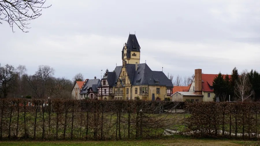 A large historical building with a tower, surrounded by trees and a hedge, under a cloudy sky.