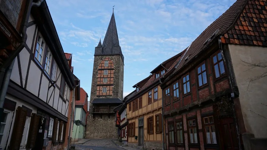 A narrow cobblestone street lined with traditional half-timbered houses leading to a tall church tower under a partly cloudy sky.