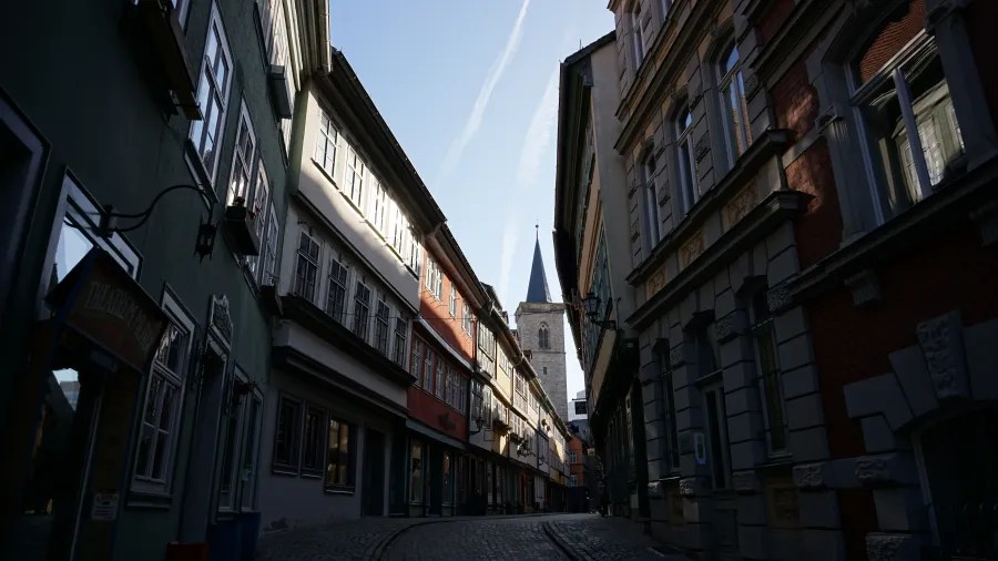 Narrow cobblestone street flanked by historic buildings, leading to a church steeple in the distance, under a clear blue sky.
