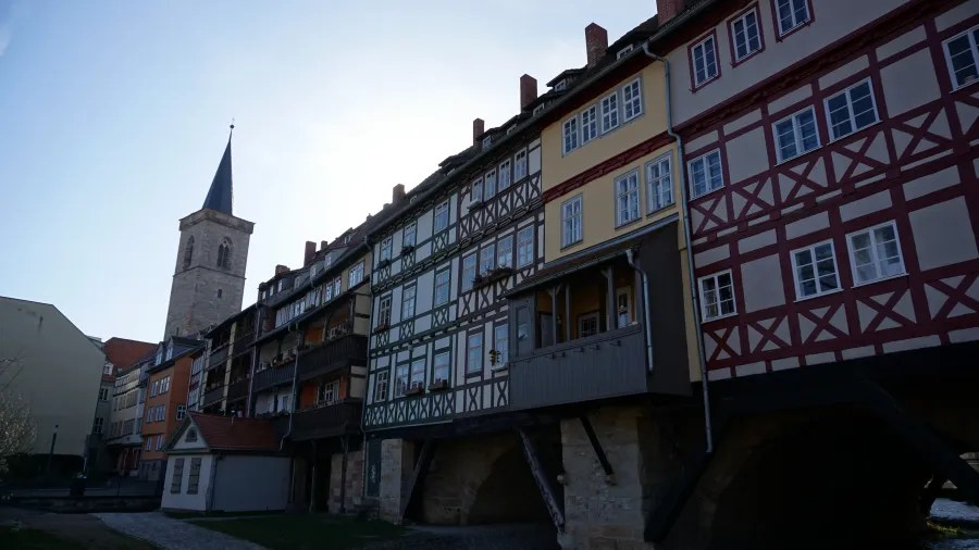 Historic timber-framed houses along the river, featuring a church steeple in the background and a clear blue sky.