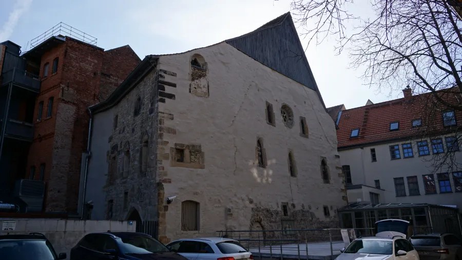 Außenansicht eines historischen Gebäudes mit restaurierter Fassade und moderner Dacheindeckung, umgeben von neueren Gebäuden und parkenden Autos.