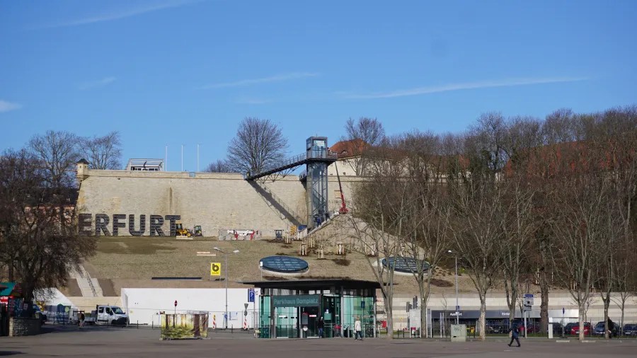 Panoramic view of Erfurt with a large wall displaying the city's name, trees lining the path, and a modern lift structure leading to the top.
