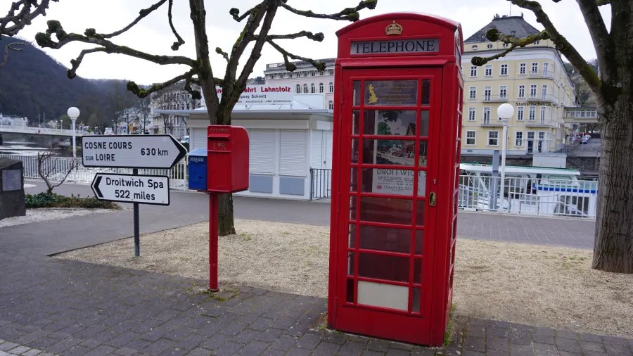 Eine rote Telefonzelle steht an einem Uferpromenade in Bad Ems, umgeben von Bäumen und Hinweisschildern, die in verschiedene Richtungen zeigen.