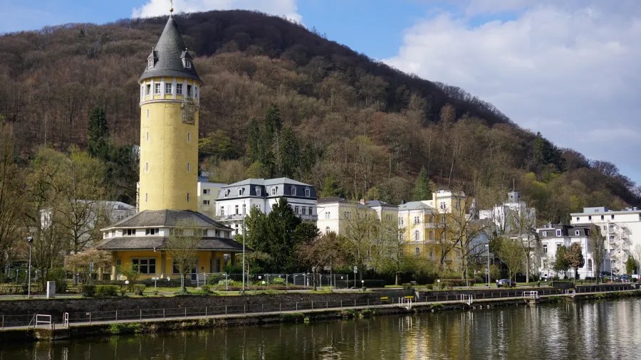 Blick auf ein historisches Gebäude mit Turm am Ufer eines Flusses, umgeben von Bäumen und Bergen im Hintergrund.