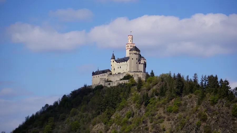 Blick auf das Schloss Stolzenfels, das auf einem Hügel über die Umgebung thront, umgeben von Bäumen und unter einem blauen Himmel mit vereinzelten Wolken.