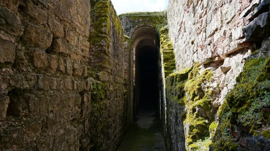 Ein schmaler, moosbedeckter Gang zwischen antiken Steinmauern, der in die Dunkelheit führt.