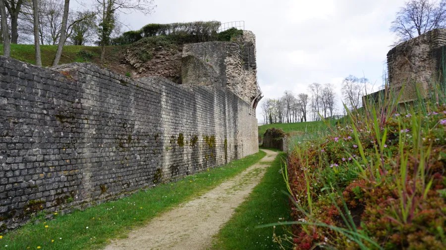 Ein gepflasterter Weg neben einer alten Steinmauer mit begrünten Bereichen und Blüten, umgeben von Bäumen und einer bewölkten Himmel.