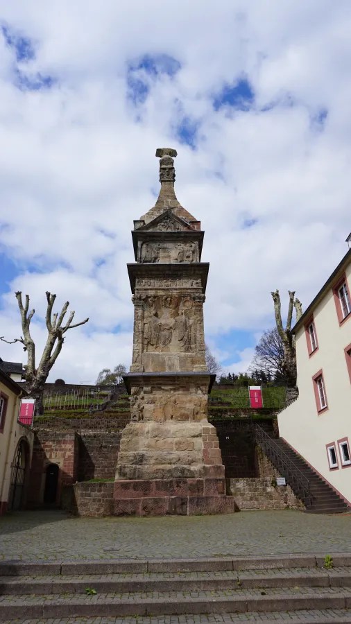 Die Igeler Säule, ein römisches Monument, erhebt sich majestätisch vor einem bewölkten Himmel. Sie zeigt kunstvolle Reliefs und steht in einer gepflasterten Umgebung mit Bäumen im Hintergrund.