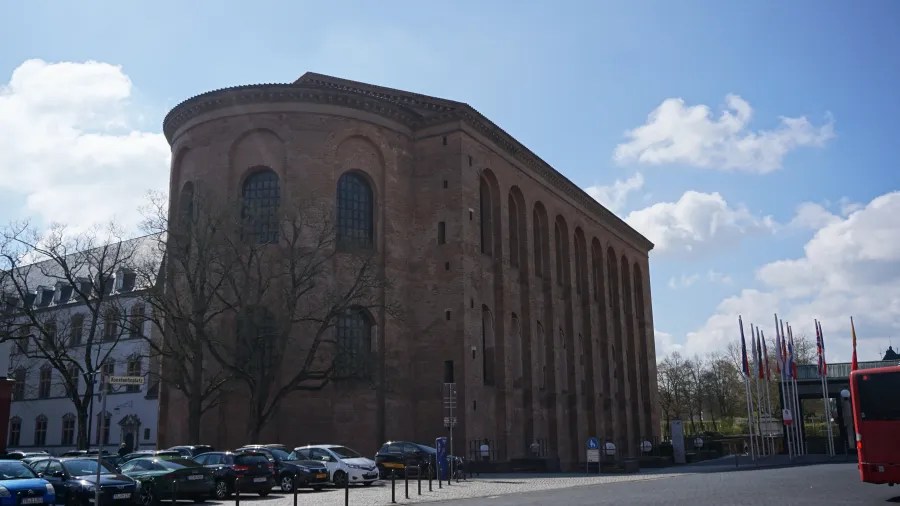 Die Konstantinsbasilika in Trier, ein beeindruckendes Gebäude aus rotem Backstein, unter einem blauen Himmel mit einigen Wolken. Vor der Basilika sind Autos geparkt, und im Hintergrund sind einige Flaggenmasten zu sehen.