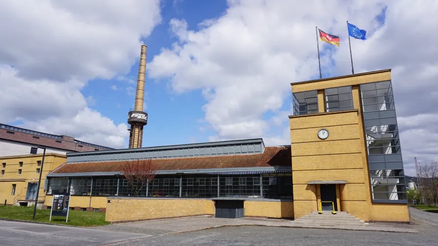 Außenansicht des Fagus-Werks mit Kamin und Flaggen, im Hintergrund bewölkter Himmel.