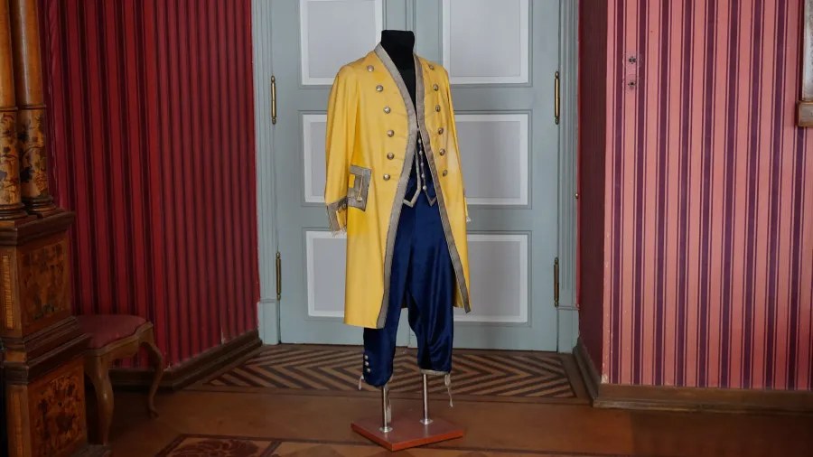 Historical outfit displayed on a mannequin, featuring a yellow coat and blue pants, set against a striped wall backdrop.