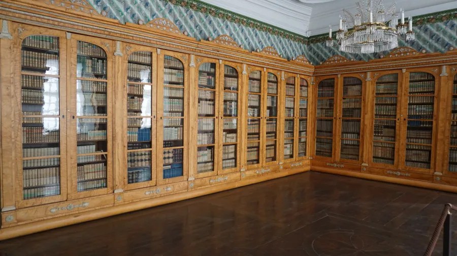A large wooden bookshelf with glass doors, filled with numerous books, located in an elegantly decorated room with a chandelier and patterned wallpaper.