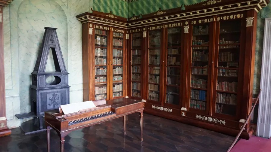 Interior of a room featuring a wooden table, a grand piano, a bookshelf filled with books, and a decorative fireplace.