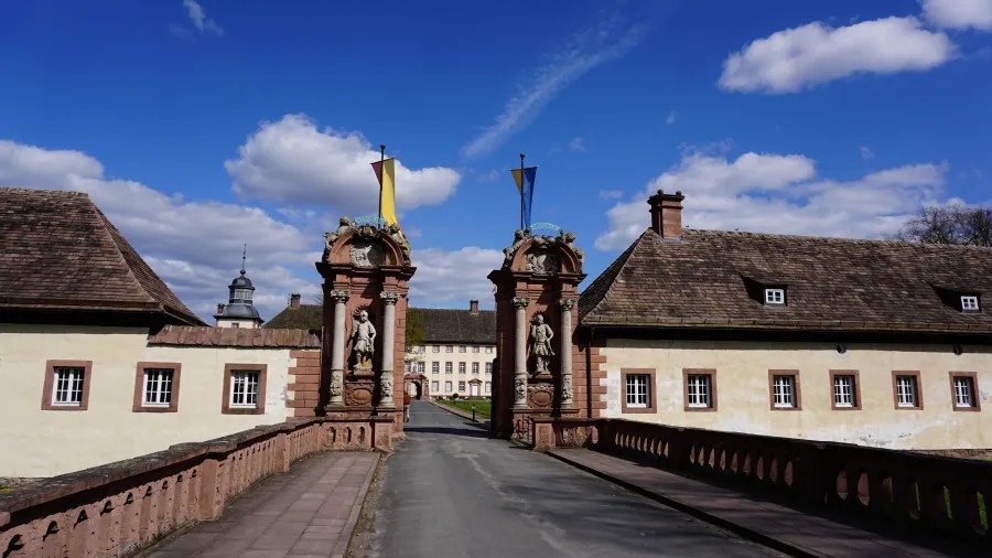 A picturesque entrance gate leading to a historic building, flanked by ornate statues and flags, under a blue sky with fluffy clouds.