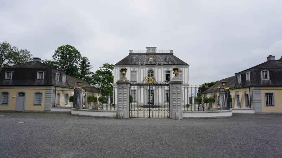 Das Jagdschloss Falkenlust in Brühl, Nordrhein-Westfalen, von der Vorderseite mit einem Zaun und Garten, umgeben von Bäumen.