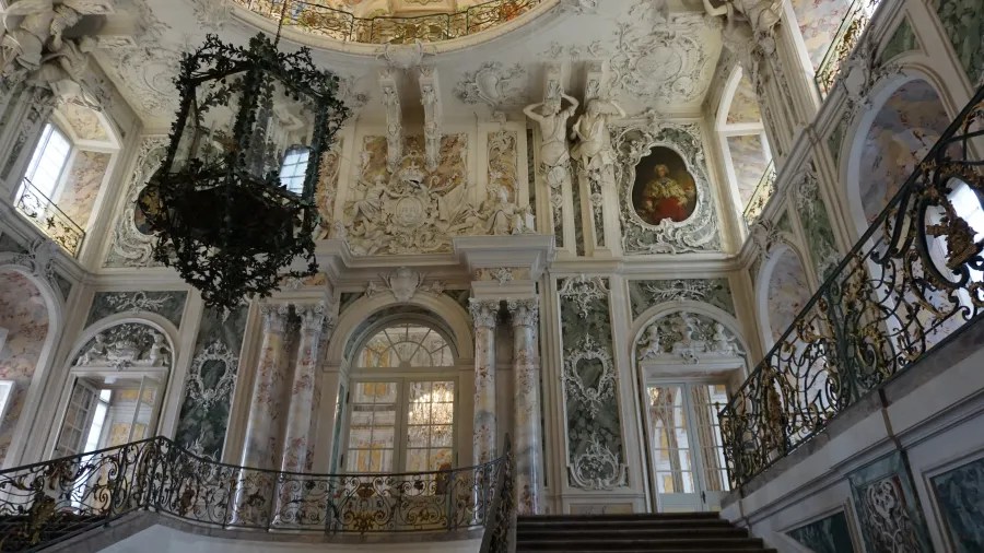 Prächtiger Innenraum eines Schlosses mit barocker Architektur, kunstvollen Wandmalereien, einem großen Kronleuchter und einer imposanten Treppe.