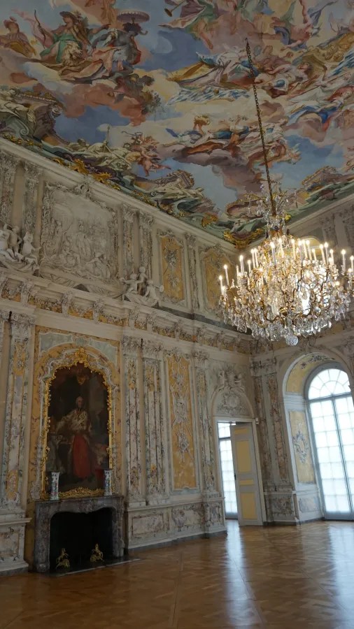 Ein opulenter Innenraum eines Schlosses mit einer kunstvollen Deckenmalerei, einem Kronleuchter und prächtigen Wänden. Die Wände sind reich verziert mit goldenen Details und Bildern.