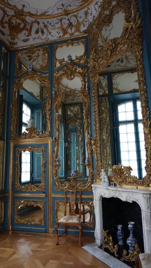 Ein prachtvoll eingerichteter Raum mit goldverzierten Spiegeln und blauen Wänden, einem Kamin aus Marmor und einem eleganten Stuhl.