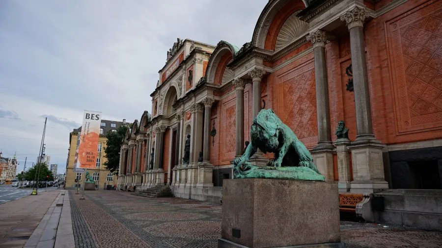 Die Ny Carlsberg Glyptotek in Kopenhagen, mit einer Statue eines Löwen im Vordergrund und rotgrauen Wänden. Die Architektur zeigt klassische Elemente und große Fenster.