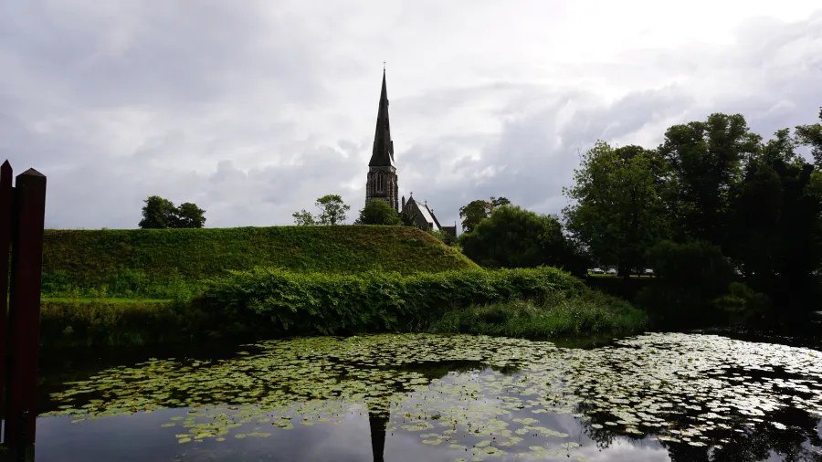 Blick auf eine Kirche mit spitzem Turm im Hintergrund, umgeben von einer grünen Wiese und einem Teich, der mit Seerosen bedeckt ist.