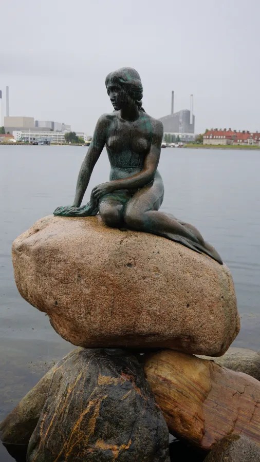 Eine Statue der kleinen Meerjungfrau auf einem Felsen im Wasser, umgeben von einer städtischen Landschaft und dunklen Wolken am Himmel.