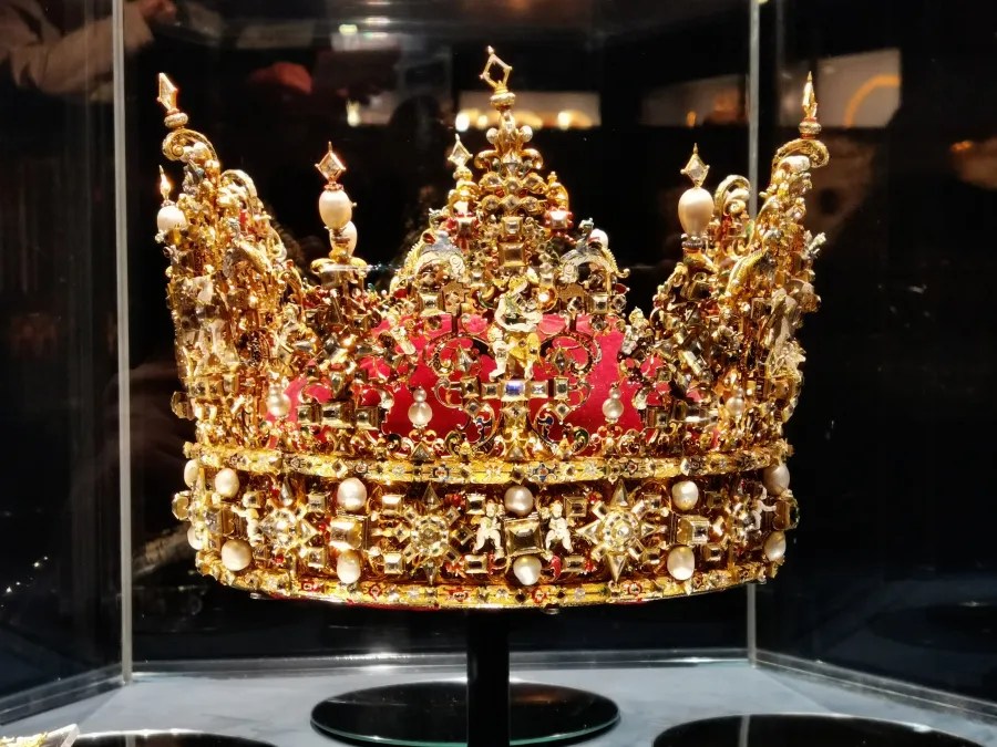 Eine kunstvoll gestaltete goldene Krone mit Perlen und Edelsteinen in einer Vitrine, umgeben von einem dunklen Hintergrund.